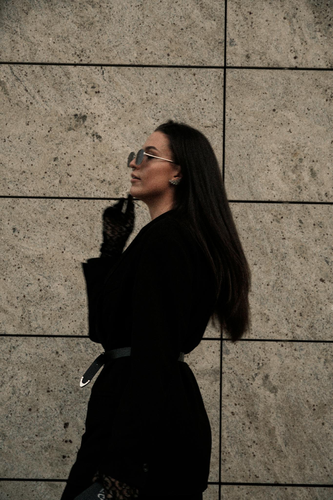 Stylish woman in black blazer posing with sunglasses against a textured wall.