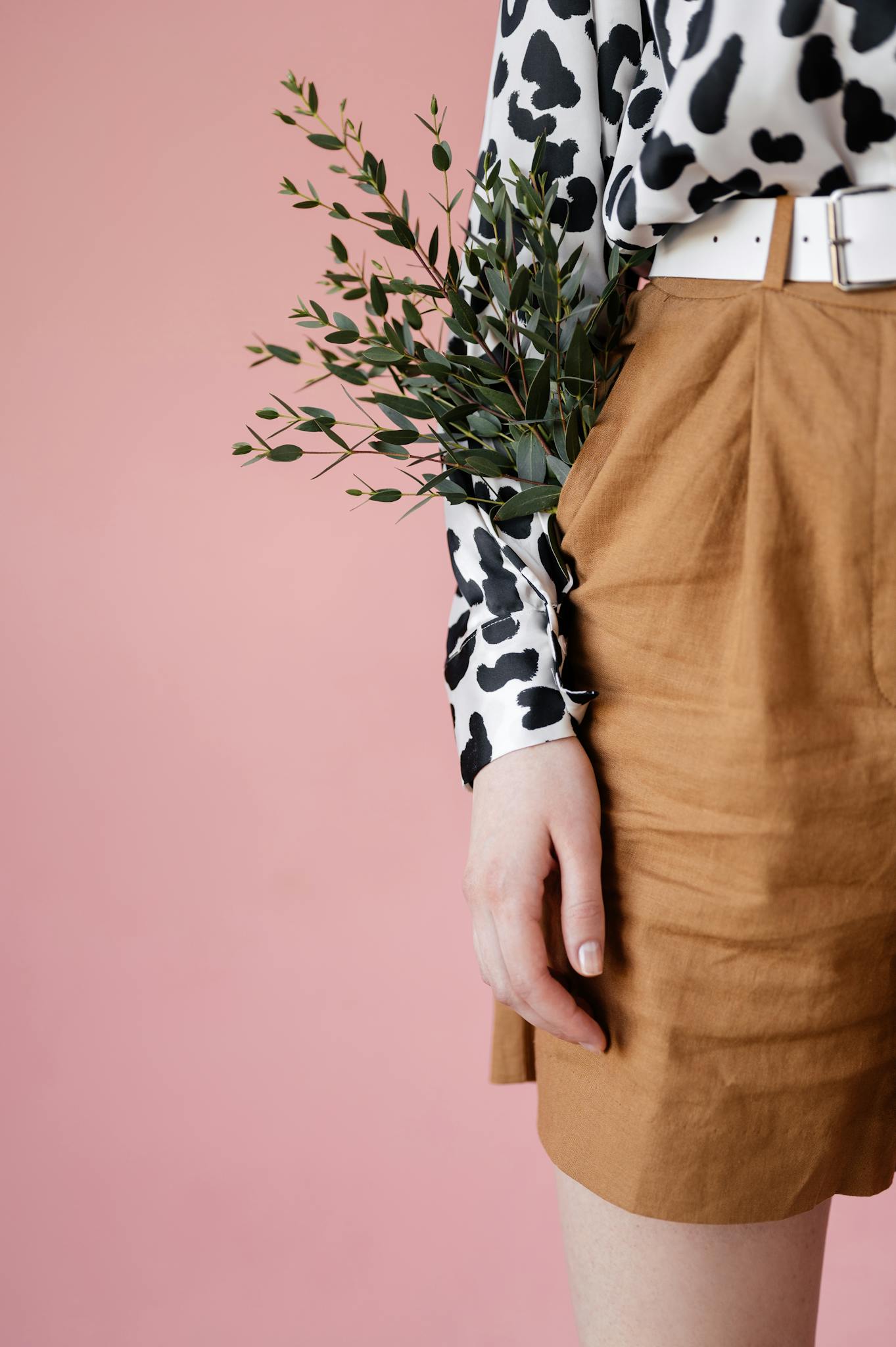 Stylish woman in patterned blouse and shorts holding greenery against a pink backdrop.