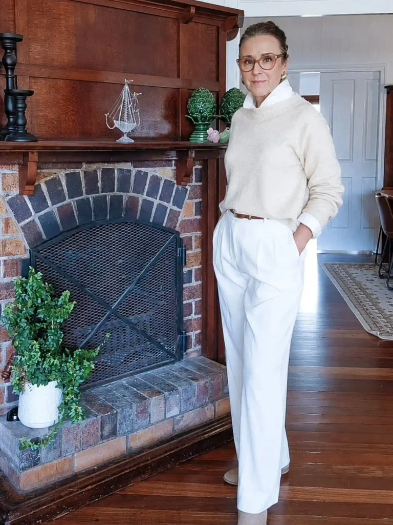 Woman over 50 in a winter white outfit with a cream sweater over a white collared shirt, pleated wide-leg trousers, and a slim brown belt