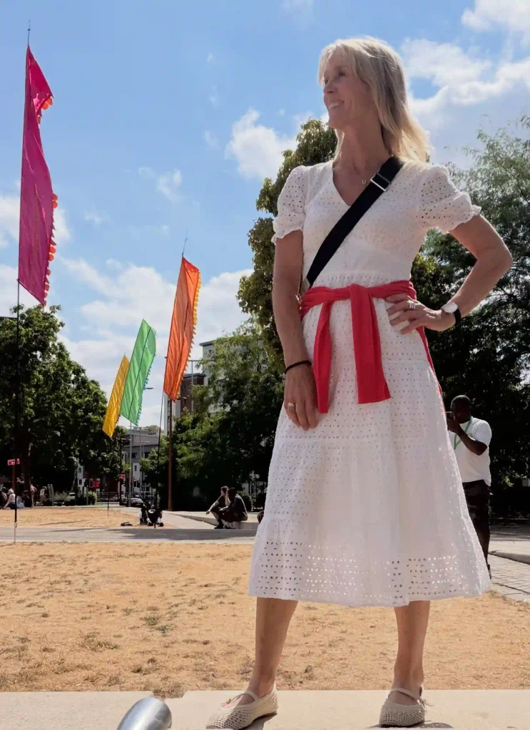 Woman in a white eyelet midi dress with a coral sweater tied at the waist, a black crossbody strap, and woven flats.