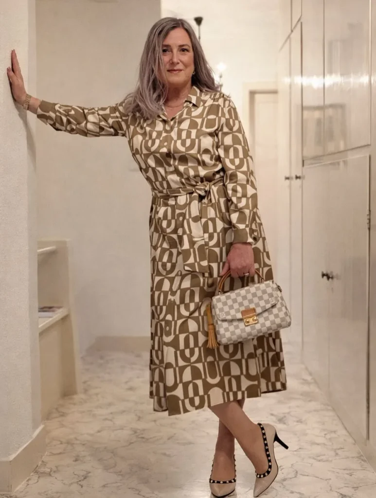 Woman with long silver hair in an olive and cream geometric shirt dress with a tie belt, beige pumps, and a small checkered top-handle bag.