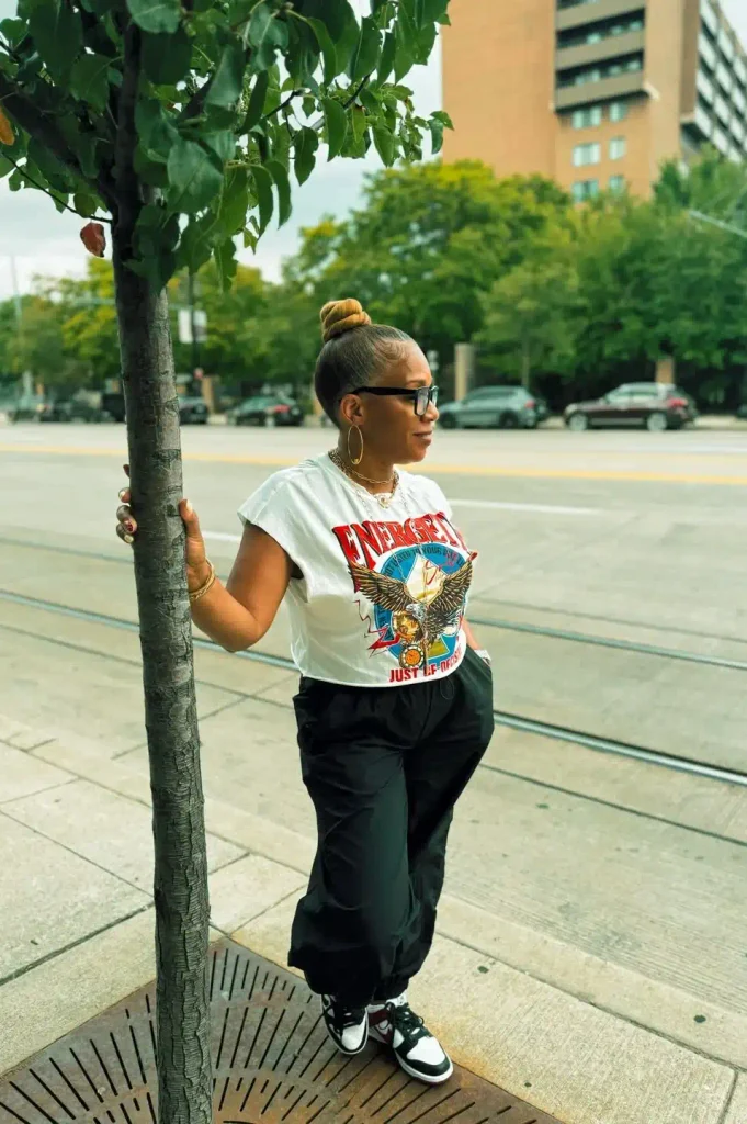 Woman with a sleek top knot in a white cropped graphic tee, black parachute pants, black and white sneakers, and gold hoops.