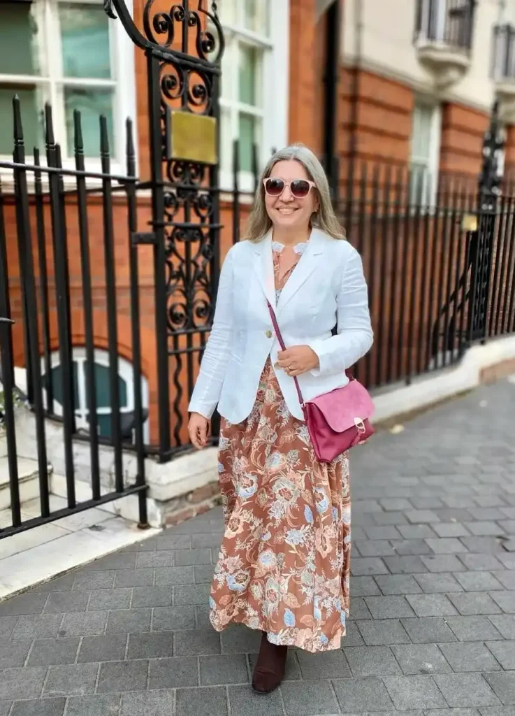 Woman with silver-blonde hair in a white linen blazer over a rust floral maxi dress, pink crossbody bag, and suede boots.