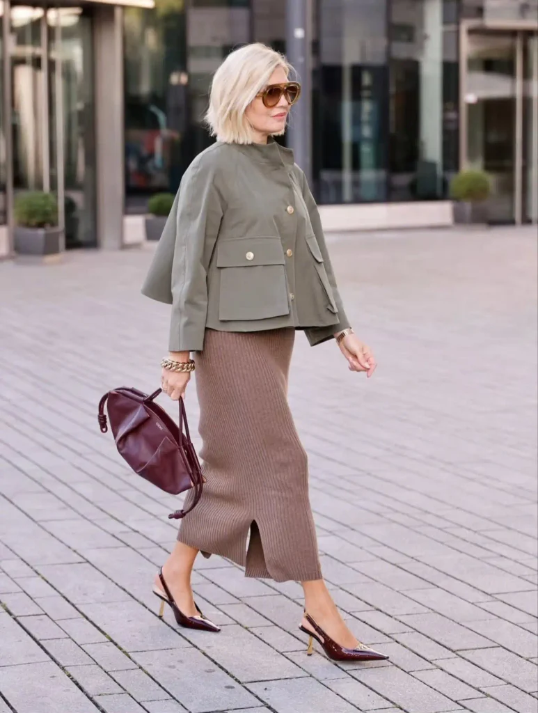 Woman with a platinum bob in an olive cropped utility jacket, mocha ribbed knit midi skirt, burgundy slingback heels, and a burgundy pouch bag.