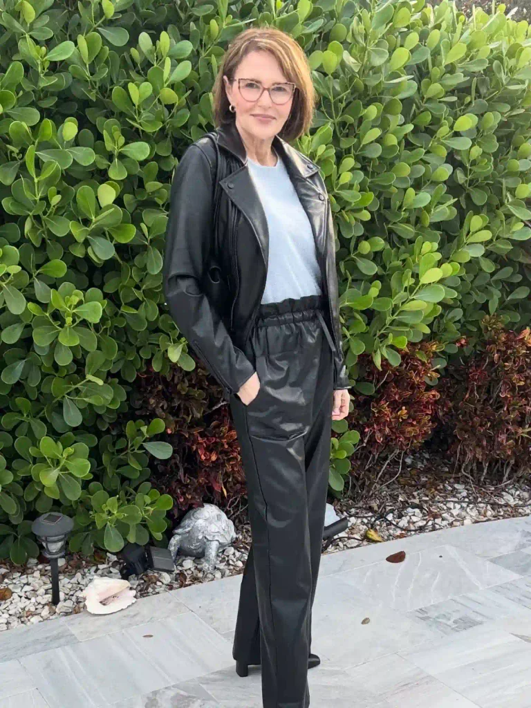 Woman in a black leather moto jacket, pale blue knit top, and black leather paperbag-waist pants in front of a green hedge
