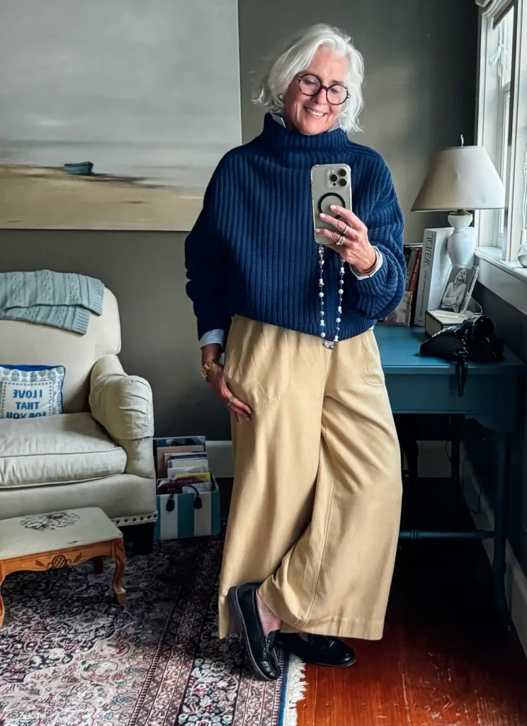 Silver-haired woman over 50 in a cropped navy knit sweater, wide-leg khaki trousers, and black loafers, taking a mirror selfie at home