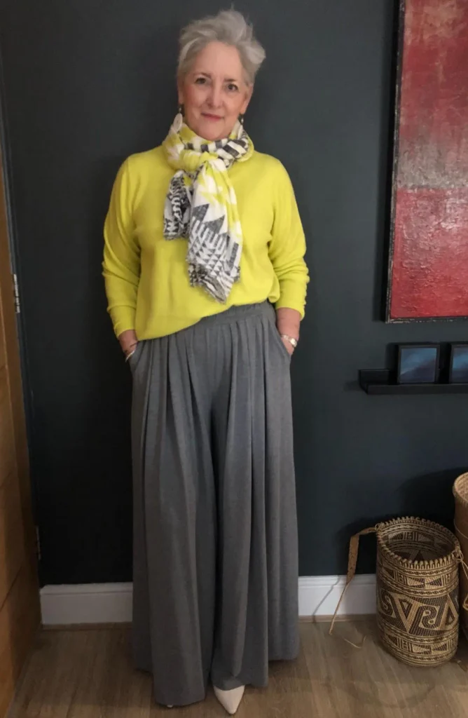 Woman with short silver hair in a bright lime sweater, geometric print scarf, wide grey jersey palazzo pants, and pointed cream shoes.