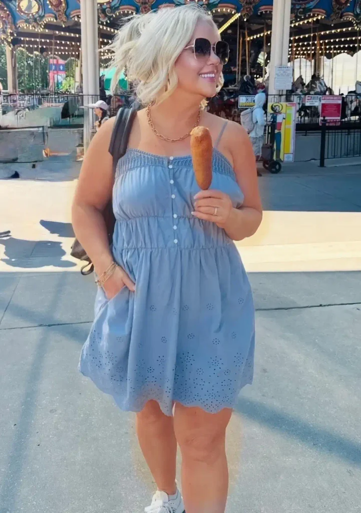 Woman holding a corn dog in a dusty blue eyelet sundress with a gold chain necklace, white sneakers, and a leather backpack.
