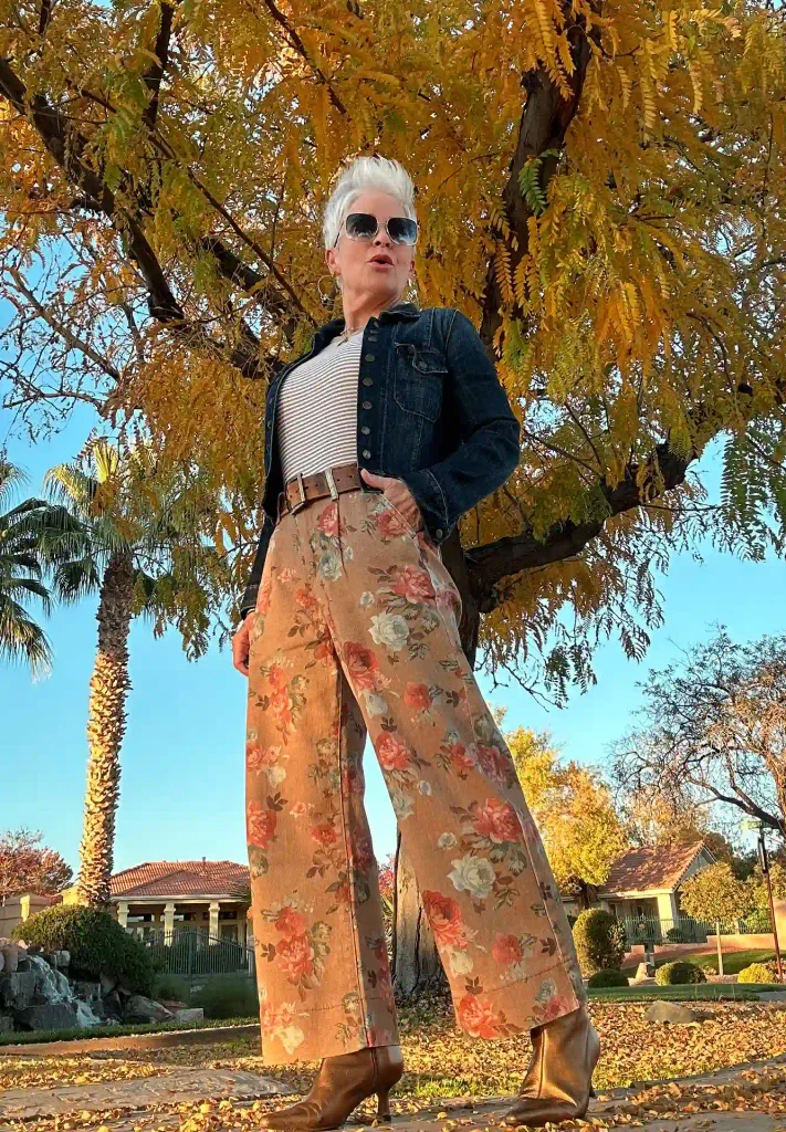 Woman over 50 with a platinum pixie in a dark denim jacket, striped tee, floral wide-leg pants, and bronze ankle boots under fall leaves.