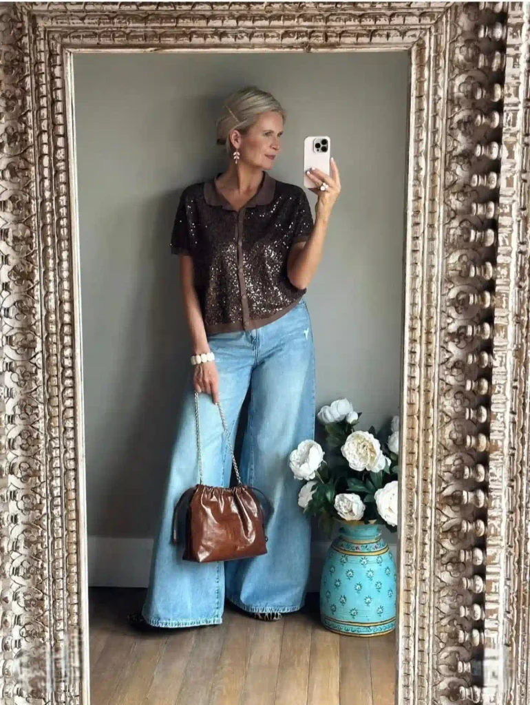 Woman in a brown sequin knit top, light wash wide-leg jeans, and leopard flats, holding a glossy brown pouch bag.